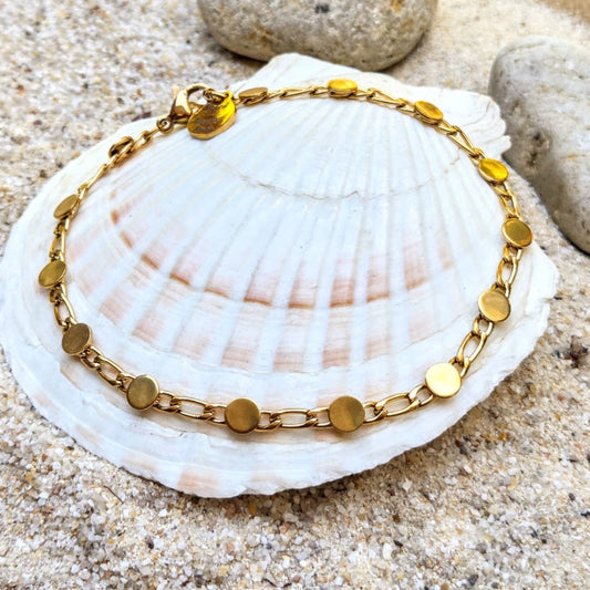 A gold chain bracelet with flat circular disc links, displayed on a white seashell over sand.