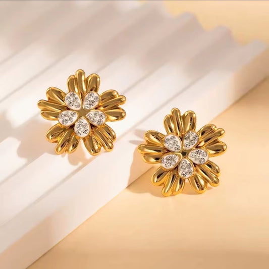 A pair of gold flower-shaped stud earrings with clear gemstones in the center, displayed on a clean white and beige background.