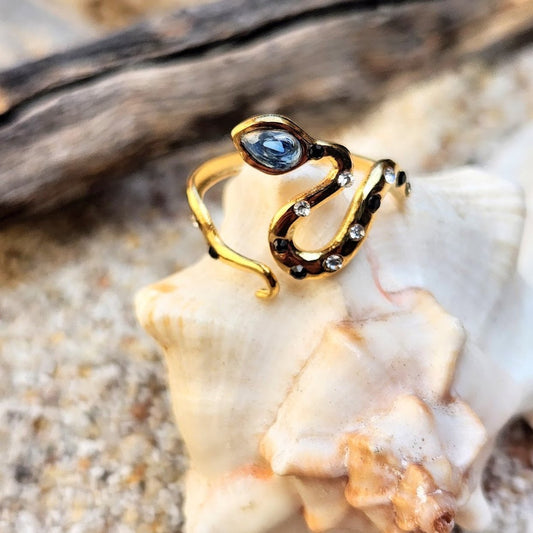 An 18k gold plated adjustable ring with blue and clear cubic zirconia layers, resembling a snake, placed on a shell on a sandy beach.