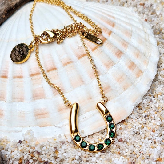 An 18k gold emerald necklace with a horseshoe pendant, featuring a gold chain and a saturn chain, placed on a shell on a sandy surface.