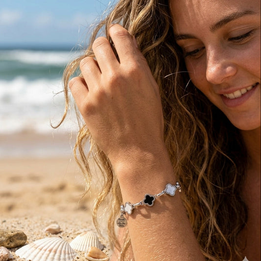 A woman on a sandy beach wearing a silver charm bracelet with black and white clover motifs. The background shows the ocean and seashells.