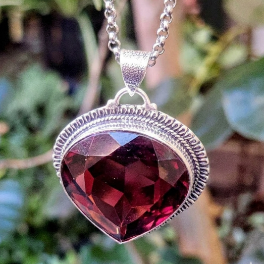A heart-shaped faceted dark red gemstone pendant in a textured silver bezel setting, hanging from a silver chain against a blurred green outdoor background.