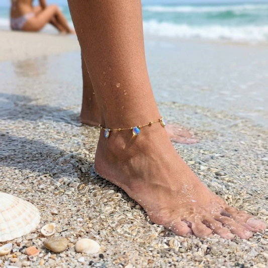 A close-up of a person's bare foot and ankle on a sandy beach, wearing a delicate gold chain anklet with small iridescent blue and white teardrop charms.