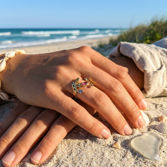 A gold ring with colorful enamel leaf details and a small clear stone, worn on a person's hand resting on a sandy beach with the ocean in the background.