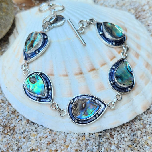 A handcrafted tear drop paua shell charm bracelet set in oxidized 925 sterling silver, displayed on a shell.