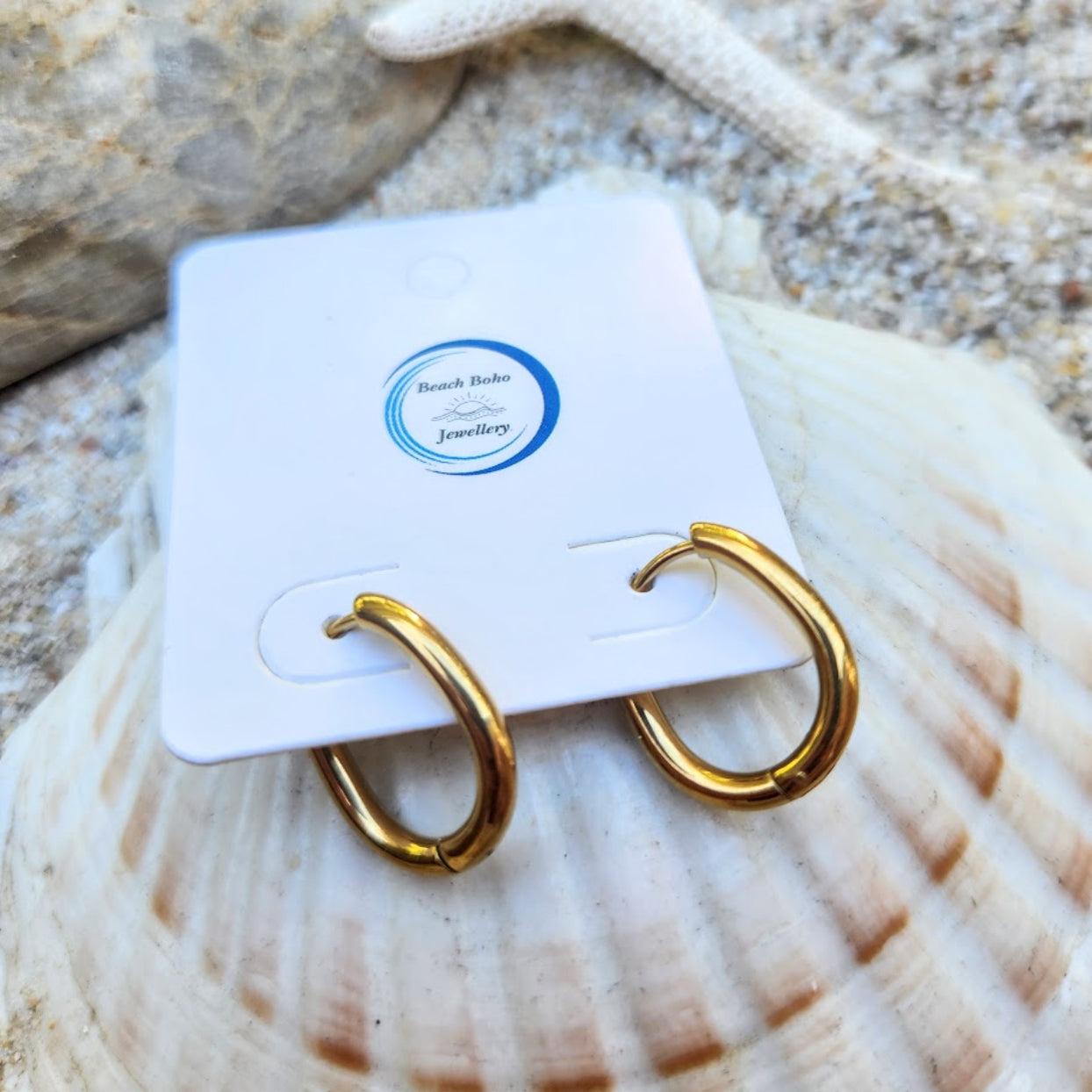 Gold hoop earrings on a white card with 'Beach Bum' branding, placed on a seashell.