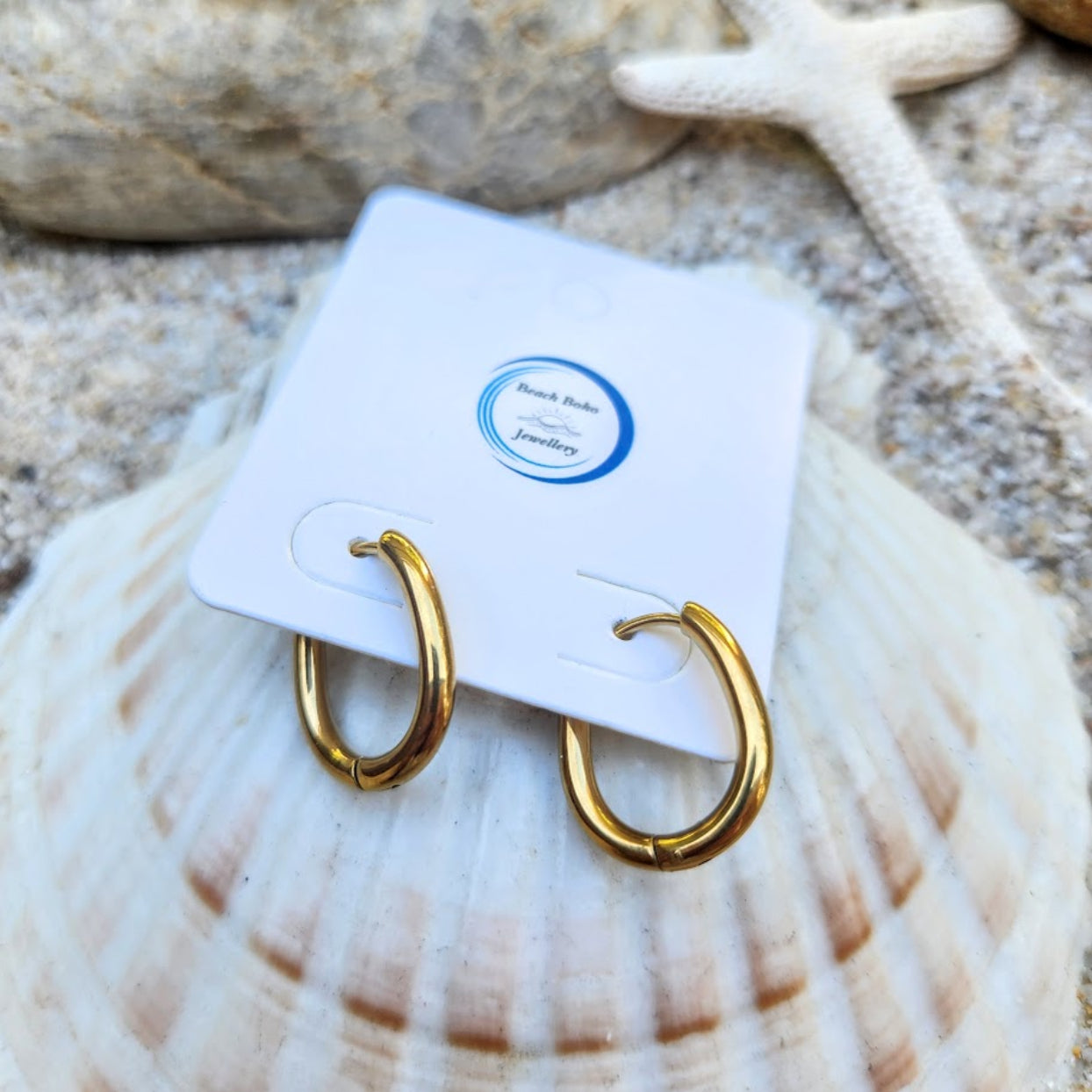 Gold hoop earrings on a white card with a blue logo, placed on a seashell.