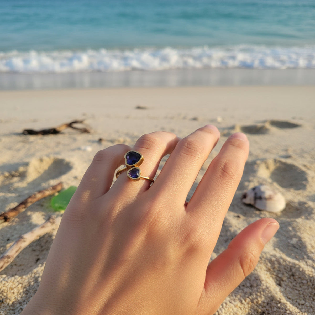 Hand wearing a gold ring with a blue stone on a sandy beach with ocean waves in the background