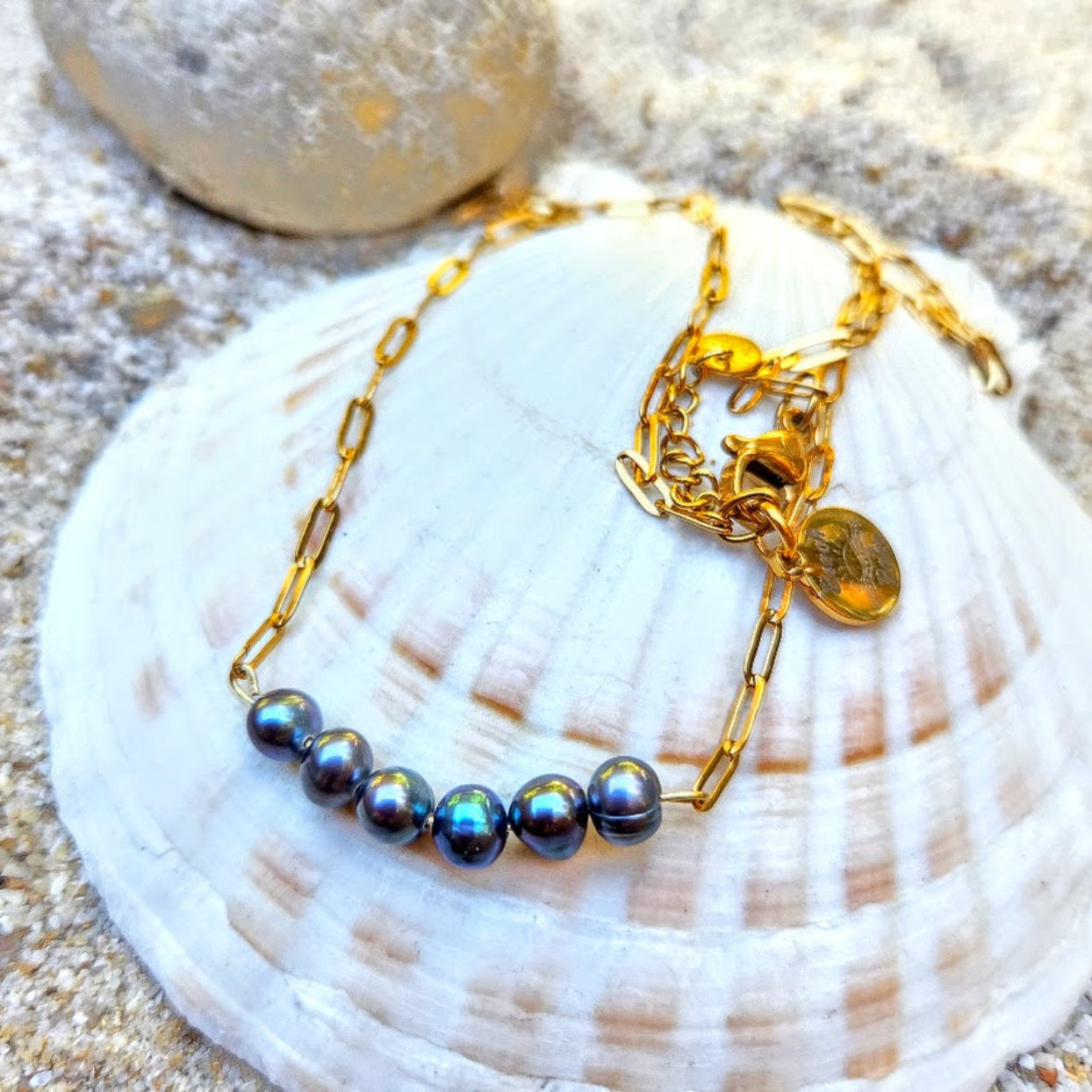 A gold chain necklace with a row of dark iridescent freshwater pearls, displayed on a white seashell over sand.