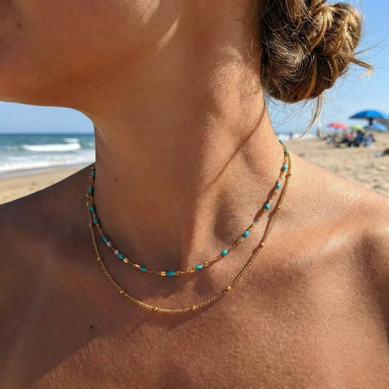 Close-up of a person wearing a gold necklace with turquoise beads on a beach.