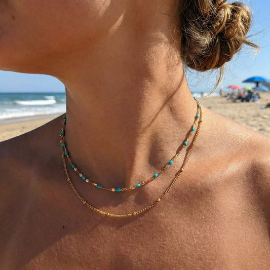 Close-up of a person wearing a gold necklace with turquoise beads on a beach.