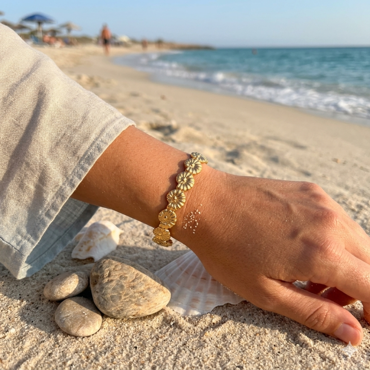 Hand wearing a gold bracelet on a sandy beach with seashells and ocean view