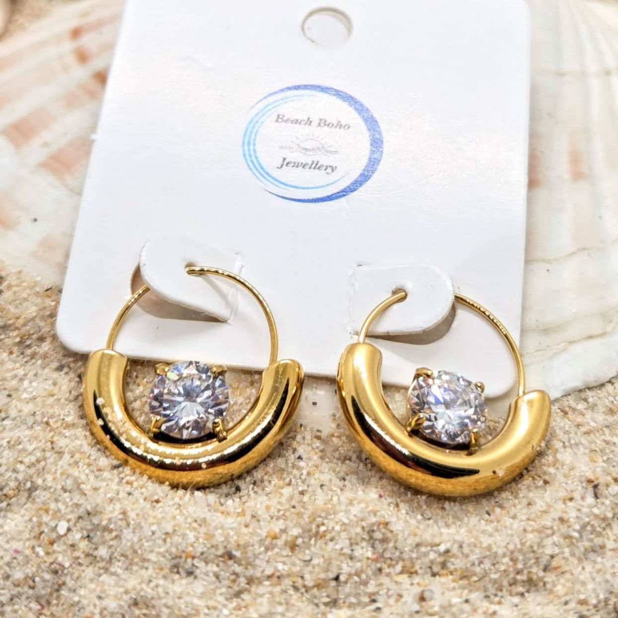 Gold hoop earrings with clear gemstones on a white card labeled 'Beach Babe Jewellery' against a sandy background.