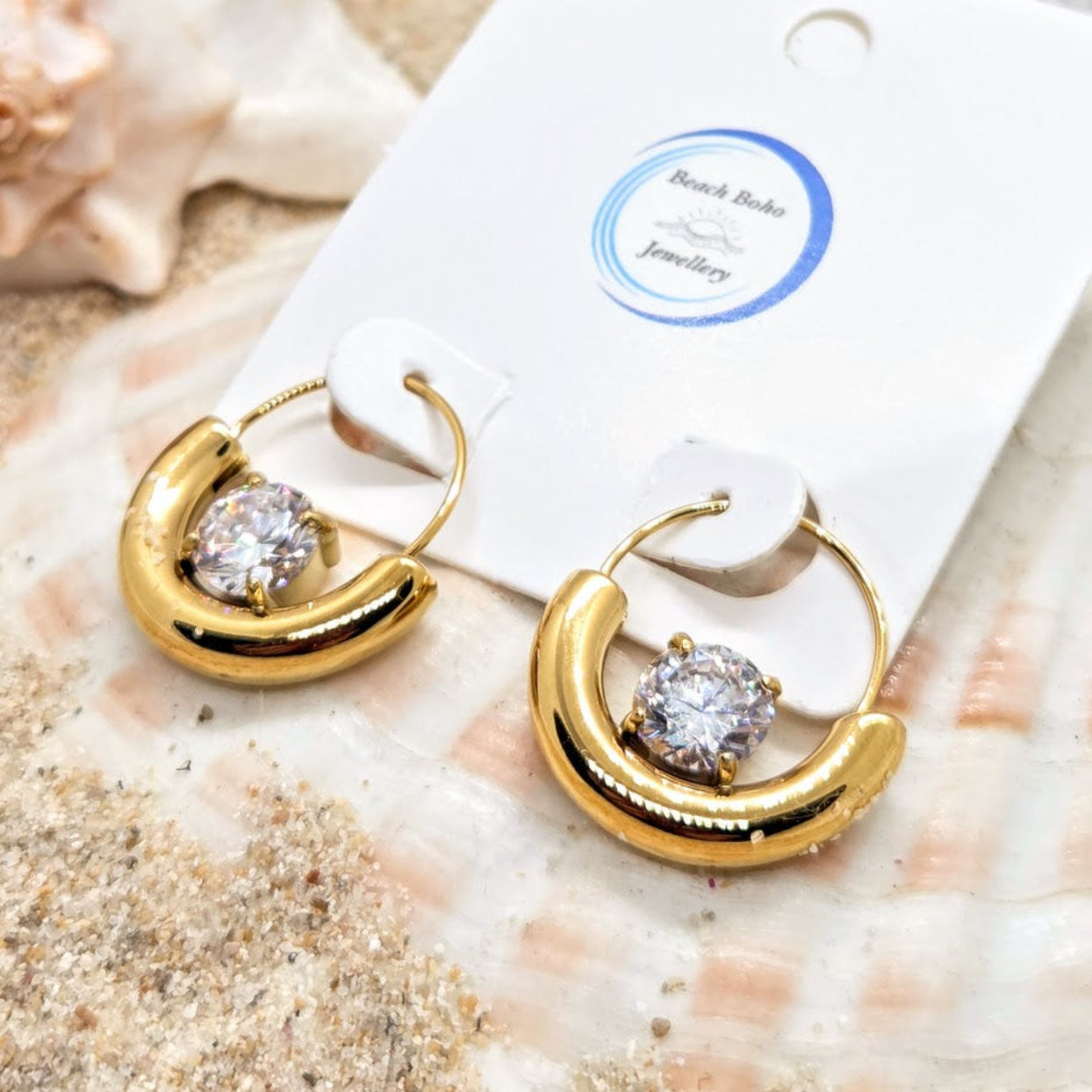 Gold hoop earrings with a central gemstone on a white card with 'Beach Babe Jewellery' label, placed on sand.