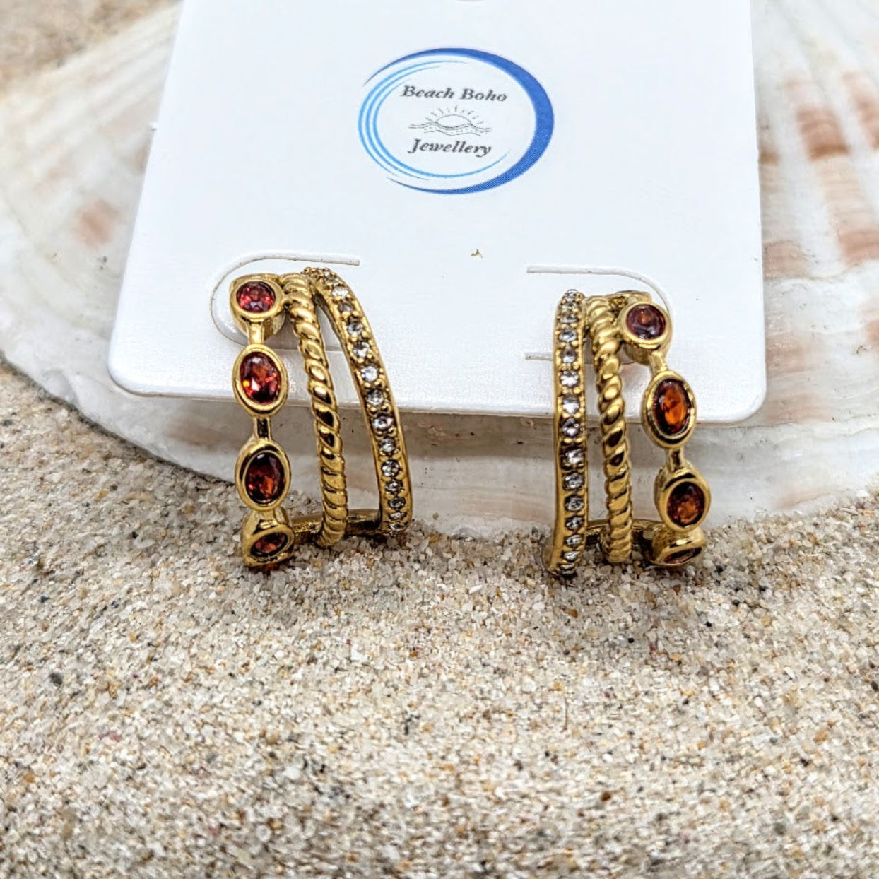 Gold hoop earrings with red gemstones on a white card labeled 'Beach Babe Jewellery' against a sandy background.