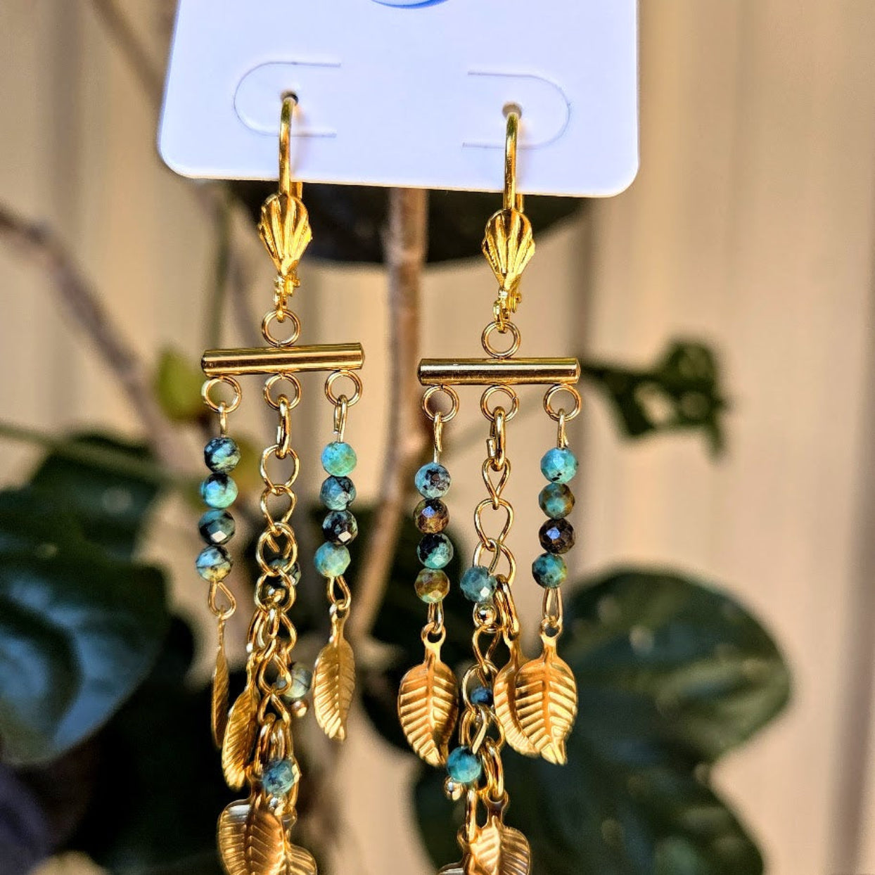 Pair of gold and blue beaded earrings on a display card with a blurred plant background.
