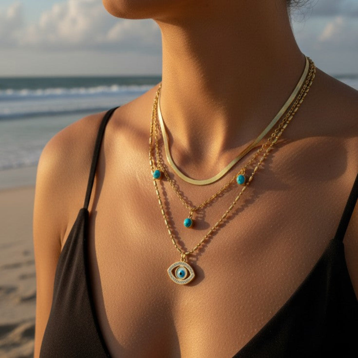 Person wearing a gold necklace with an eye pendant on a beach