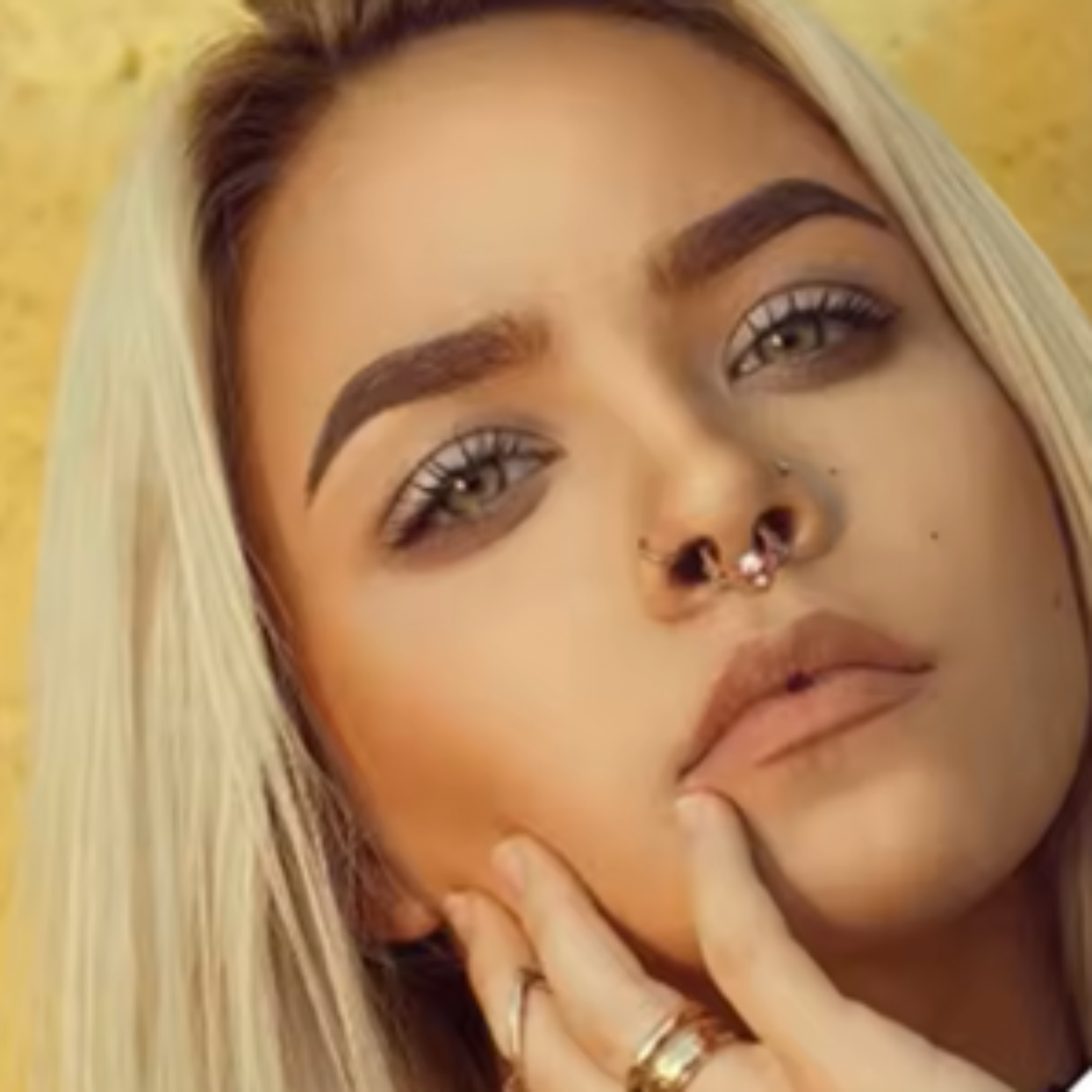 Close-up of a person with blonde hair and piercings, wearing gold jewelry against a yellow background.