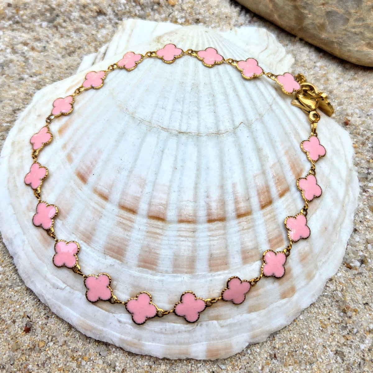 A gold chain bracelet with pink clover-shaped links, displayed on a large white seashell over sand.