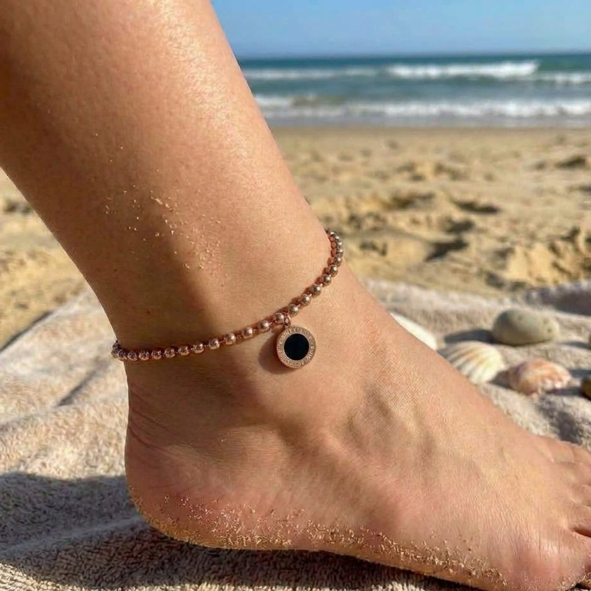 A delicate rose gold beaded anklet with a central black and rose gold circular charm is shown on a person's ankle, standing on a towel at a sandy beach with the ocean in the background.
