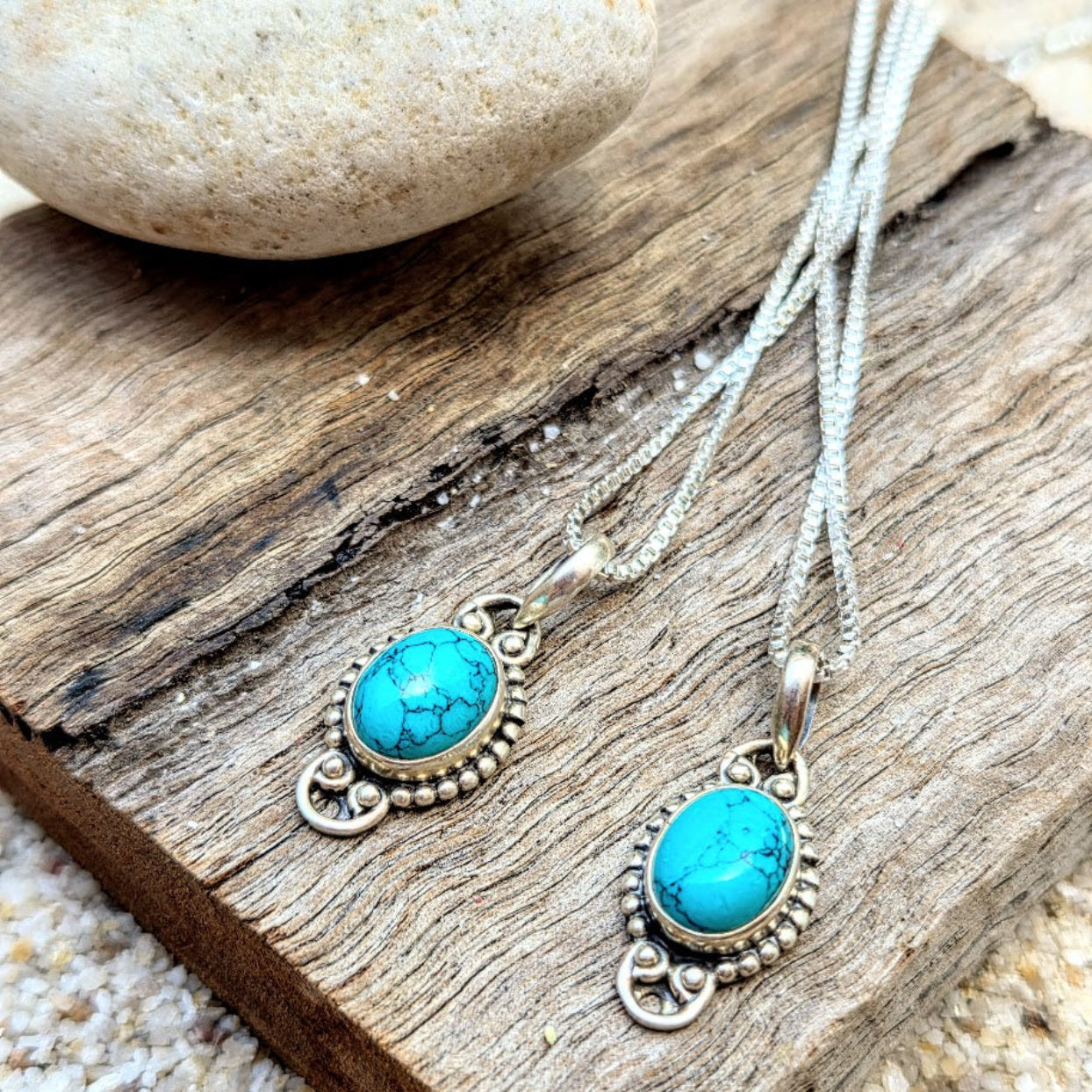 Turquoise stone pendants on silver chains on a wooden surface with pebbles.