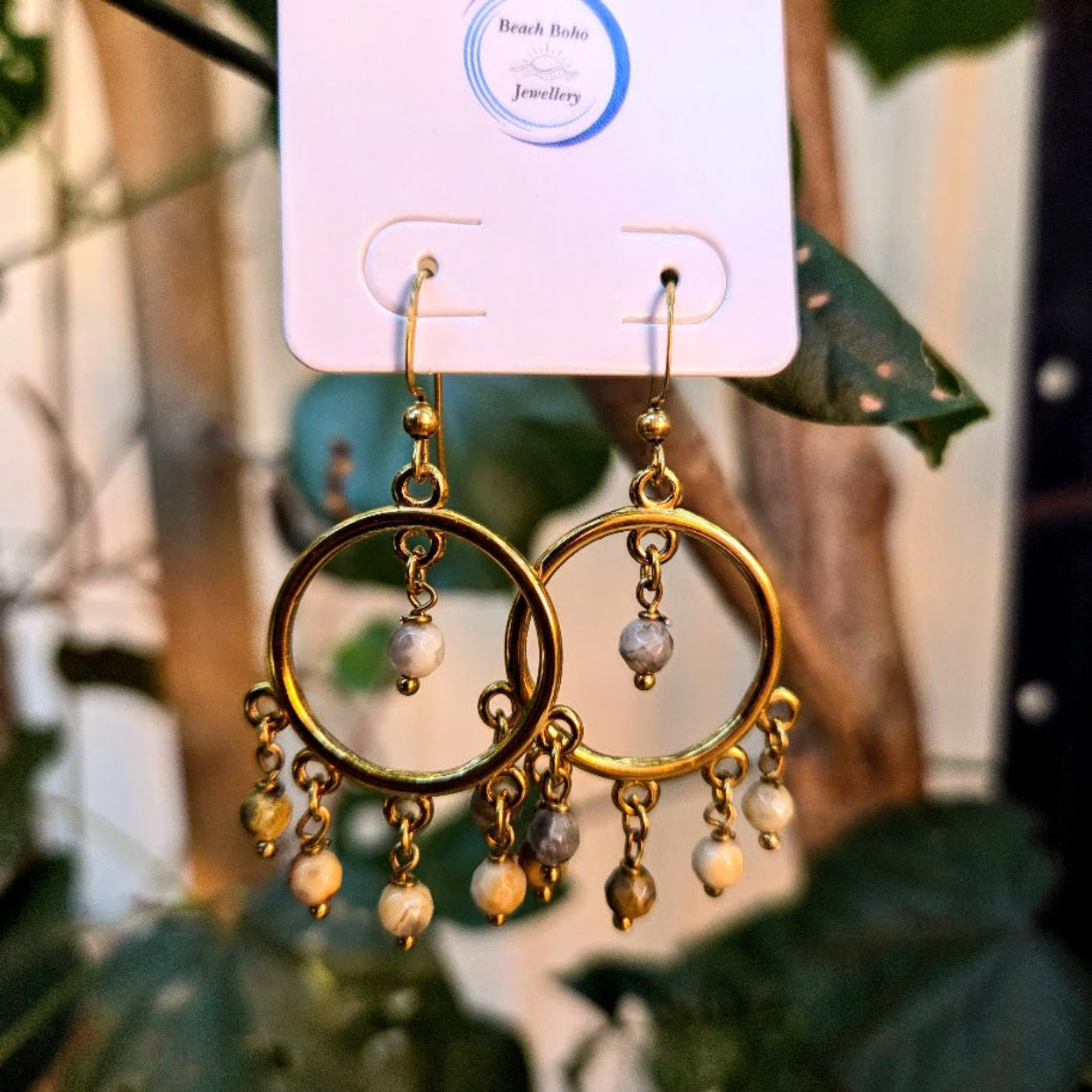 Gold hoop earrings with pearl-like beads on a branded card, blurred green leaves in the background
