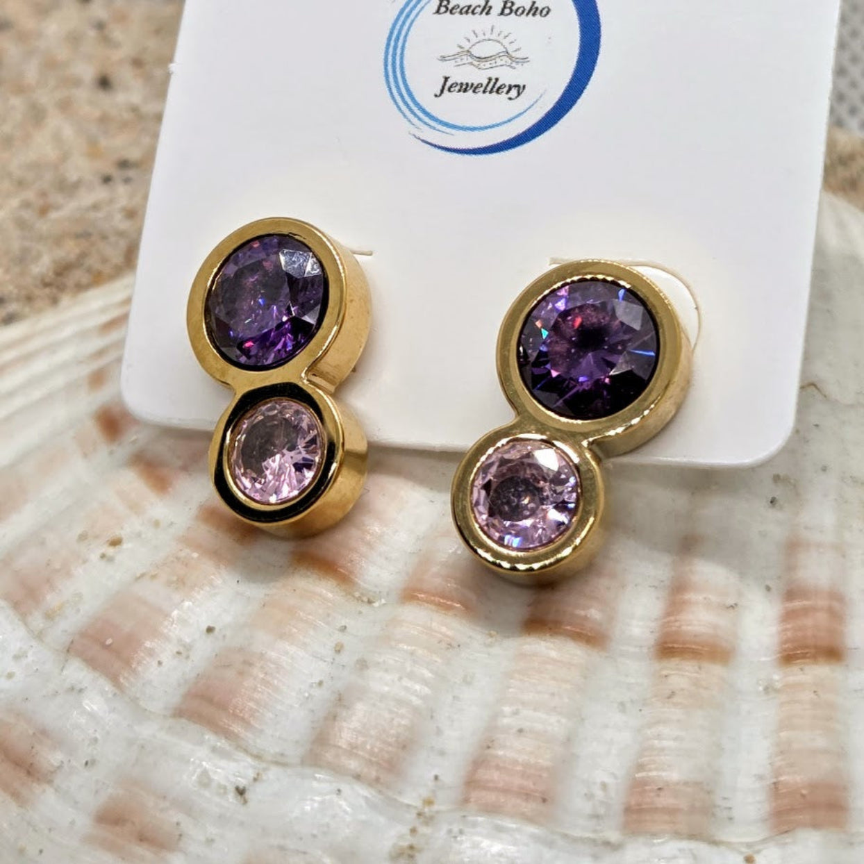 Gold earrings with purple and pink gemstones on a 'Beach Boho Jewellery' card over a shell background.