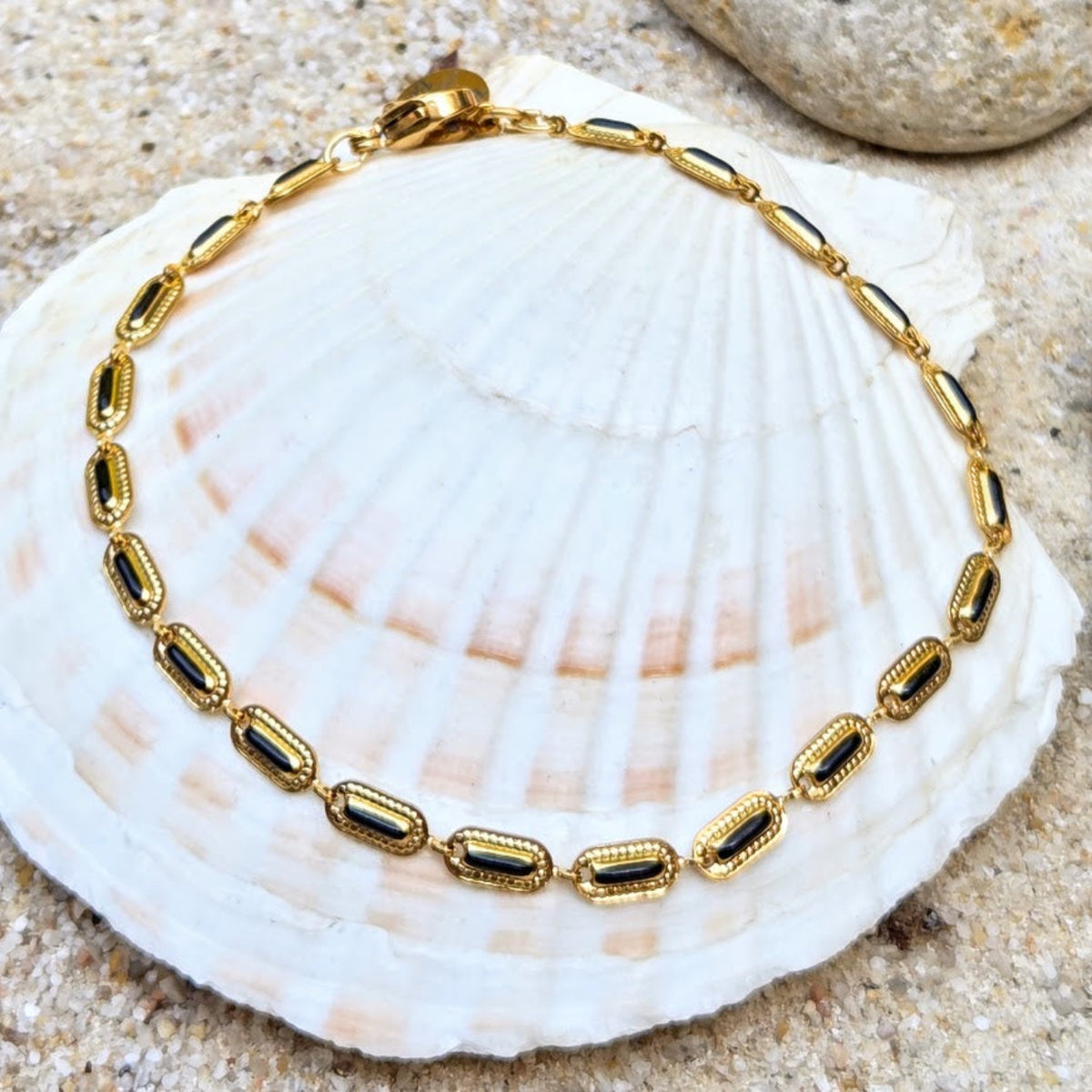 A gold chain bracelet with rectangular black inlay stones, displayed on a large white seashell over sandy ground with a rock in the background.