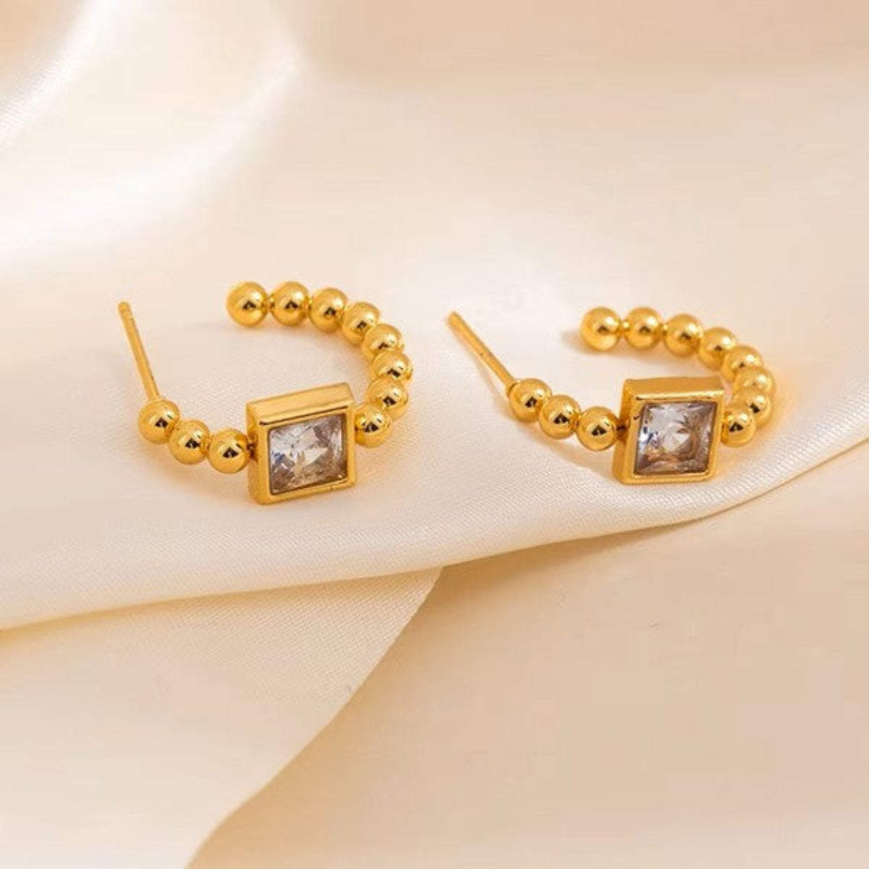 A pair of gold hoop-style earrings with small gold beads and a square clear gemstone in the center, displayed on a soft cream satin fabric background.
