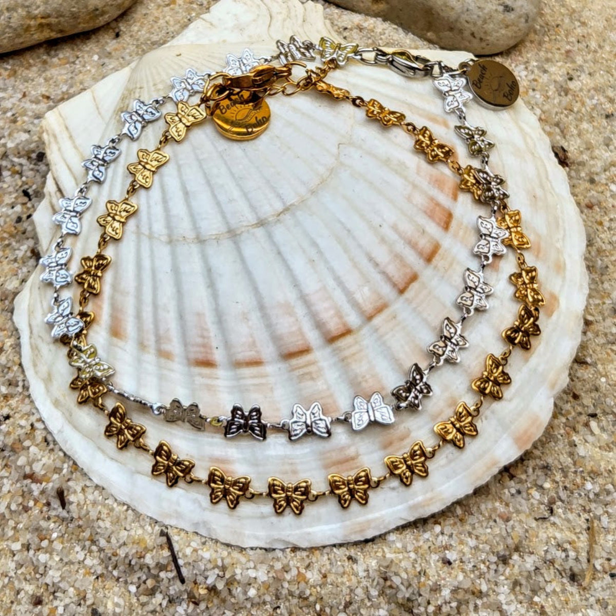 A double-strand butterfly charm bracelet in gold and silver-tone metal, displayed on a large white seashell over a sandy beach background.