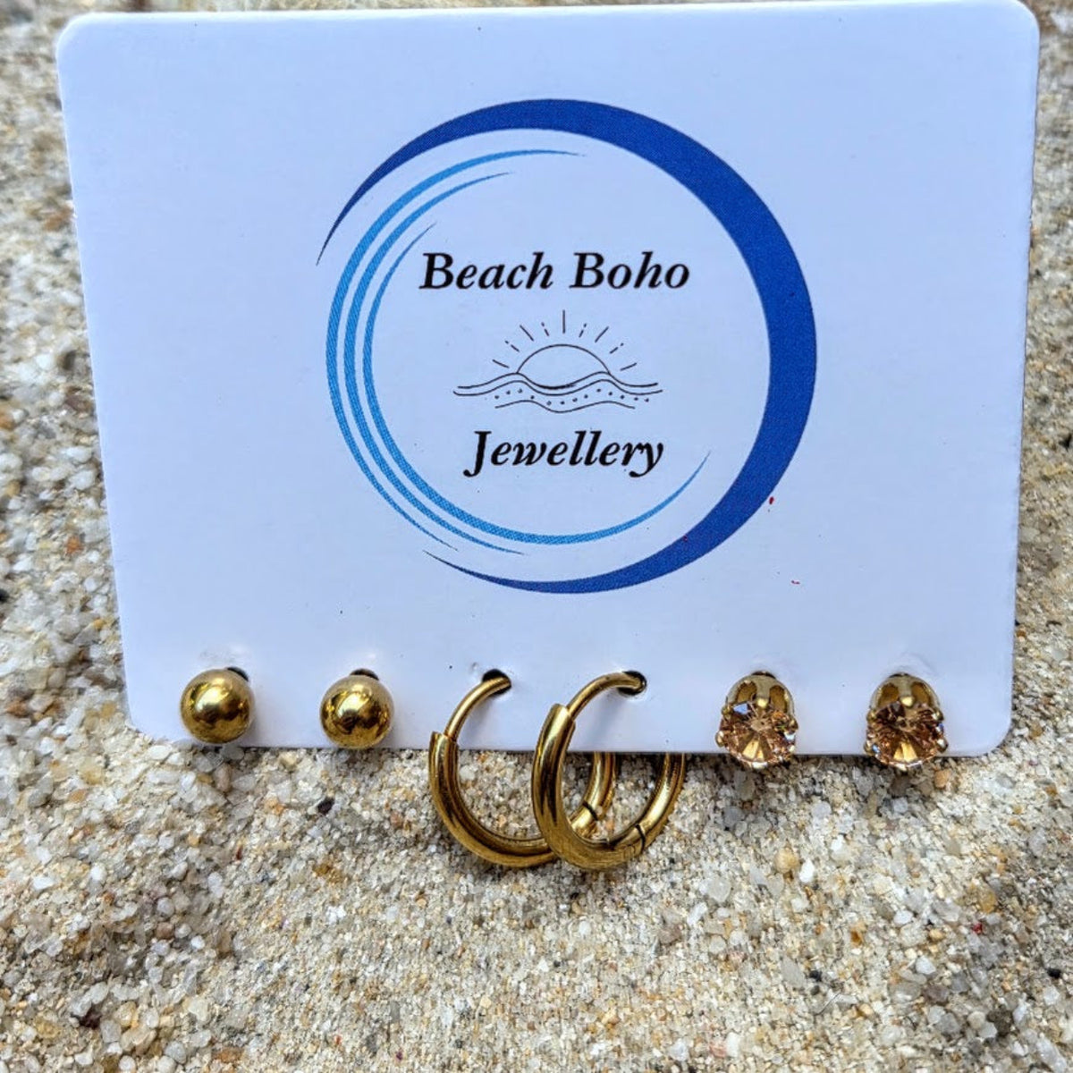 Gold hoop earrings and stud earrings displayed on a white card branded 'Beach Boho Jewellery', set on sandy ground.