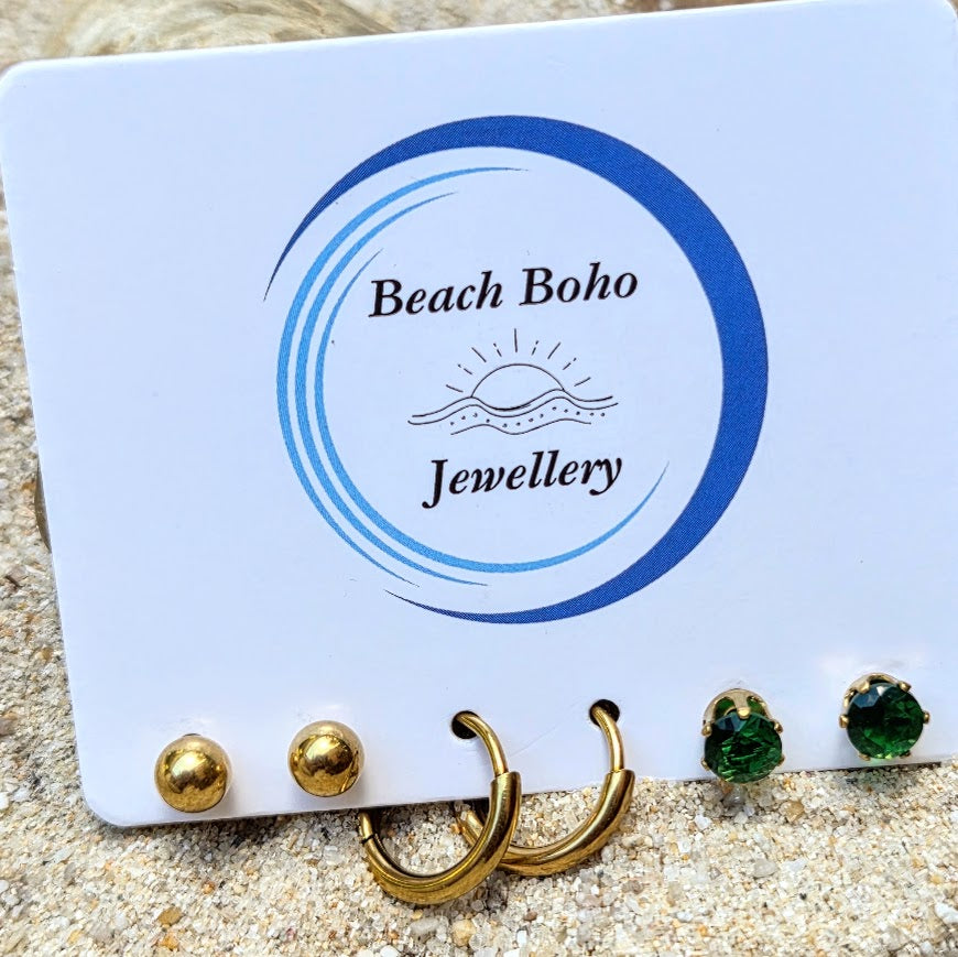 A white display card branded 'Beach Boho Jewellery' holding several earrings on a sandy surface. The earrings include two gold studs, two gold hoop earrings, and two green gemstone stud earrings.