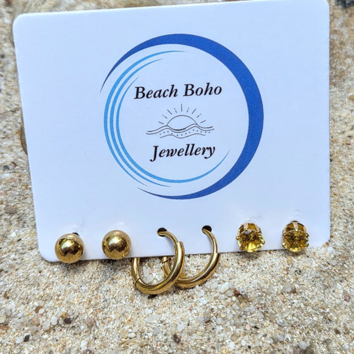 A set of gold-toned earrings displayed on a white card labeled Beach Boho Jewellery, placed on sandy ground. The earrings include three gold ball studs, two gold hoop earrings, and two gold oval stud earrings with yellow gemstones.