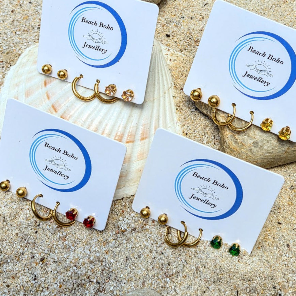 Four pairs of gold hoop earrings displayed on white jewelry cards with the brand name 'Beach Boho Jewellery' printed on them. The earrings feature various accents including small gold beads, clear crystals, red and green stones, and one gold hoop with a yellow gem.