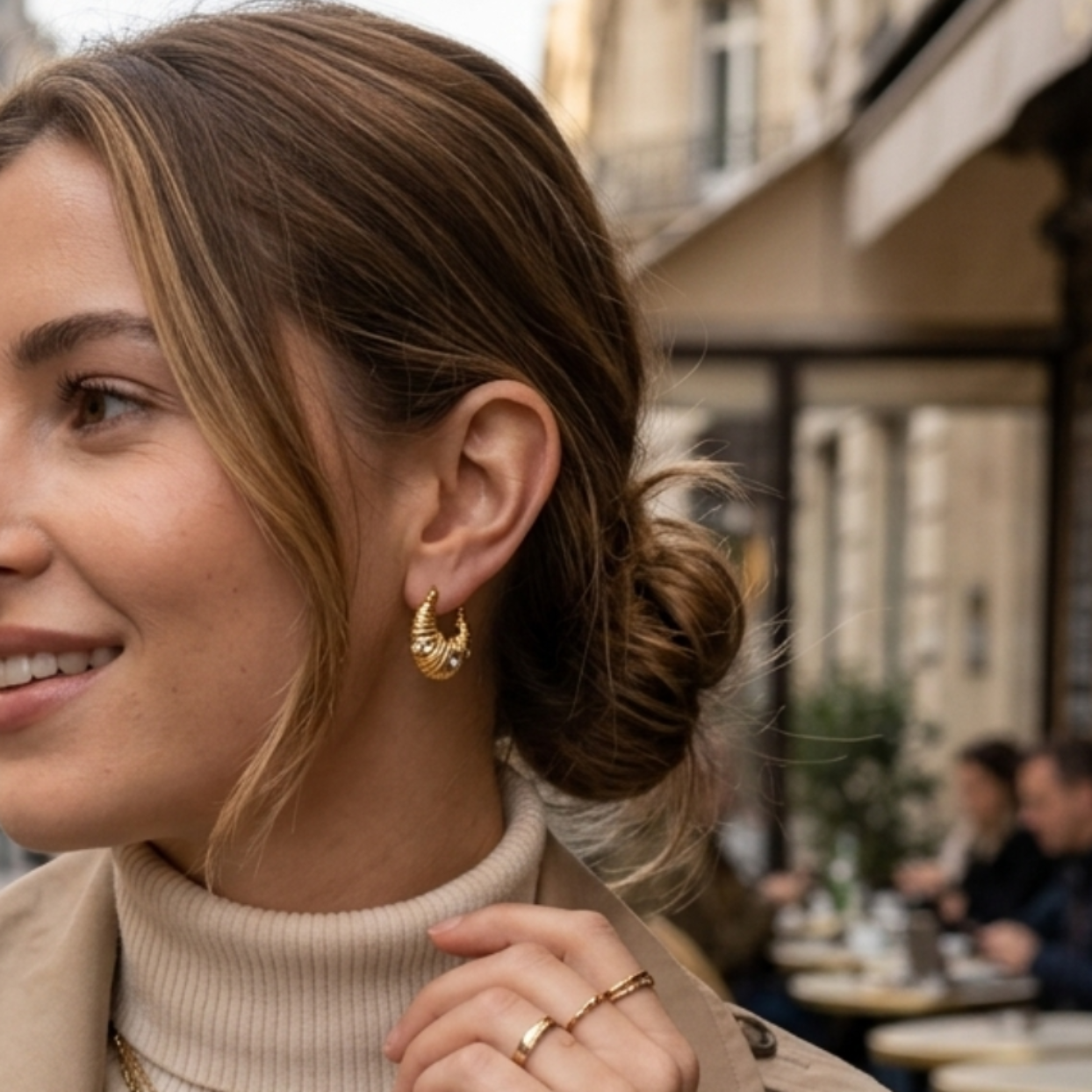 A woman wearing a beige trench coat over a cream ribbed turtleneck, accessorized with layered gold necklaces, gold hoop earrings, and a gold watch, standing on a city street.