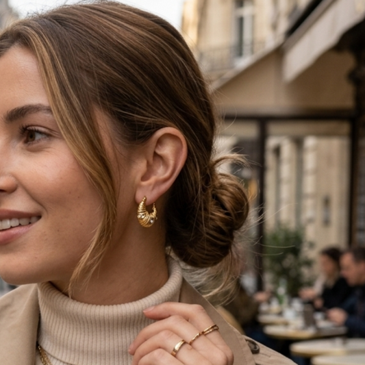 A woman wearing a beige trench coat over a cream ribbed turtleneck, accessorized with layered gold necklaces, gold hoop earrings, and a gold watch, standing on a city street.