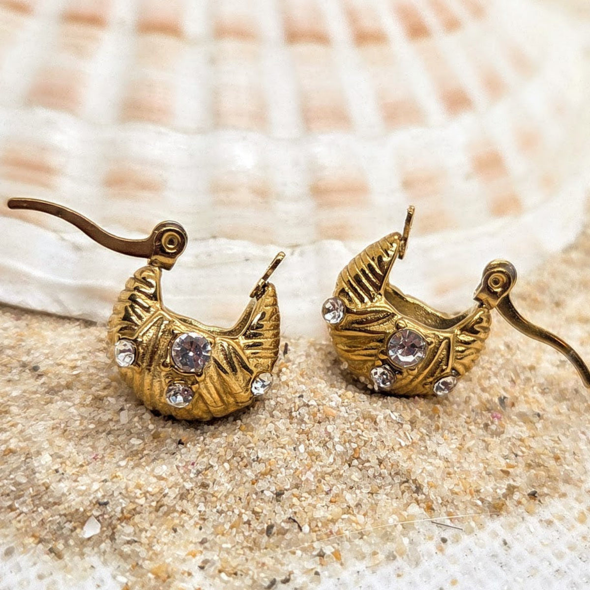 A pair of gold crescent moon earrings with textured detailing, clear rhinestones, and hook backings, displayed on sand in front of a seashell.