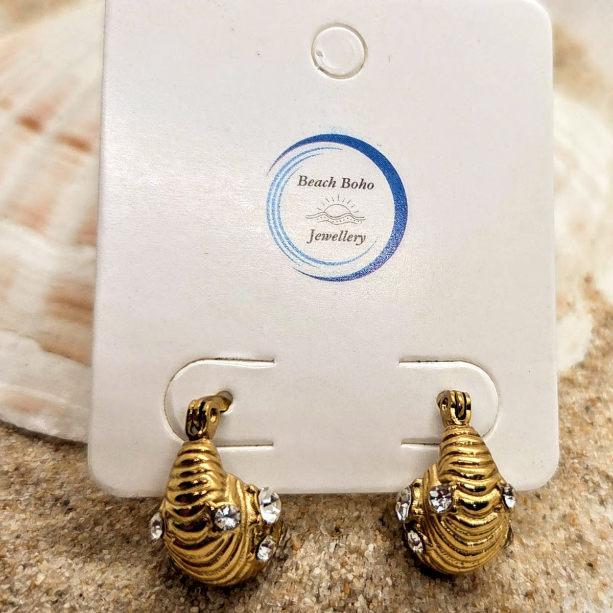 A pair of gold-toned shell-shaped hoop earrings with small clear rhinestones, displayed on a white jewelry card labeled Beach Boho Jewellery, set against a sandy, seashell-background.