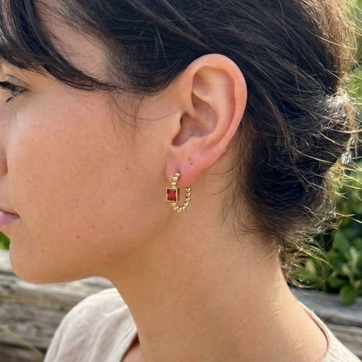 A woman in profile wearing a small gold hoop earring with a red-orange stone, photographed outdoors near beach grass and shells.