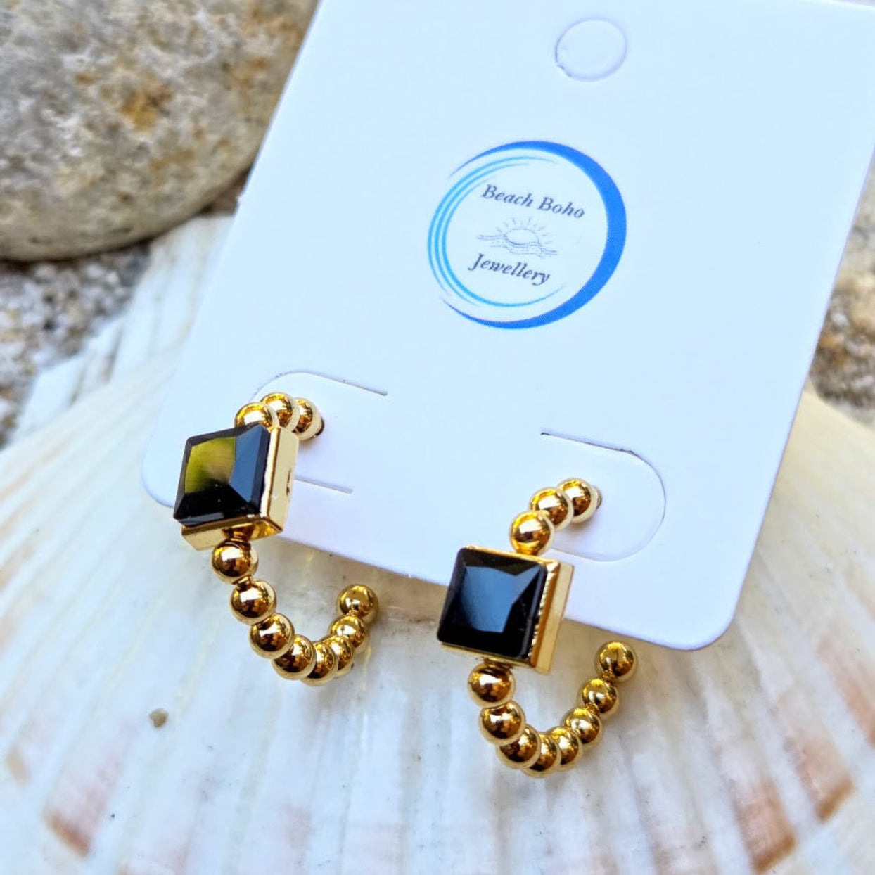 A pair of gold huggie hoop earrings with square black gemstone accents, displayed on a white Beach Boho Jewellery card resting on a seashell.
