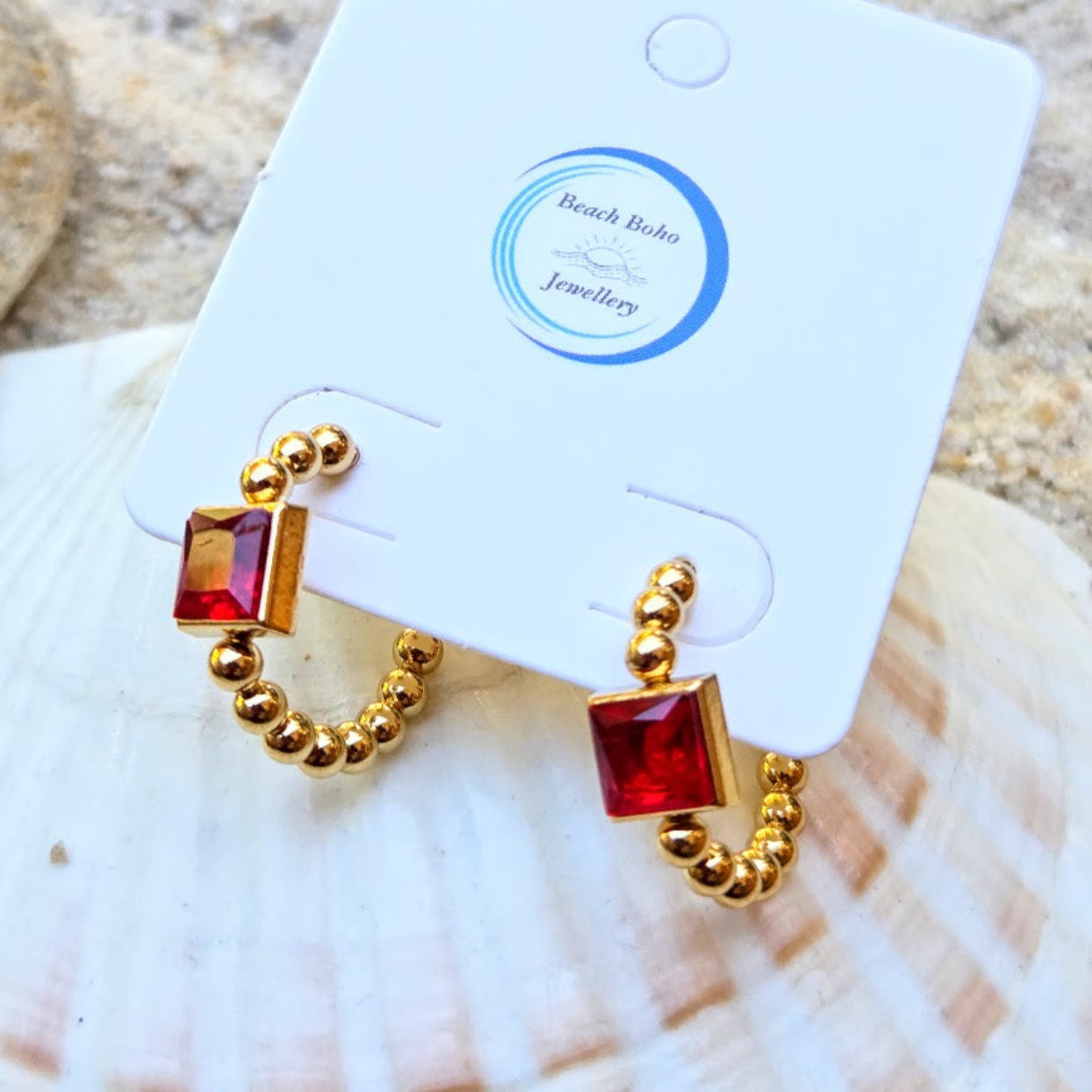 A pair of gold hoop earrings with square red gemstones, displayed on a white card labeled Beach Boho Jewellery, resting on a seashell.