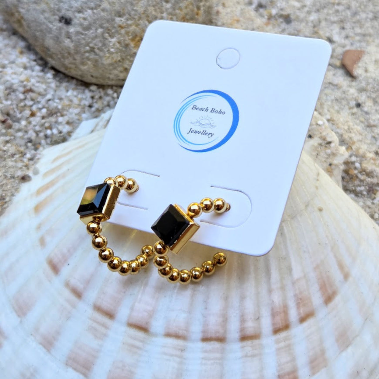 A pair of gold crescent hoop earrings with black square stones, displayed on a white card labeled 'Beach Bibo Jewellery' resting on a seashell.