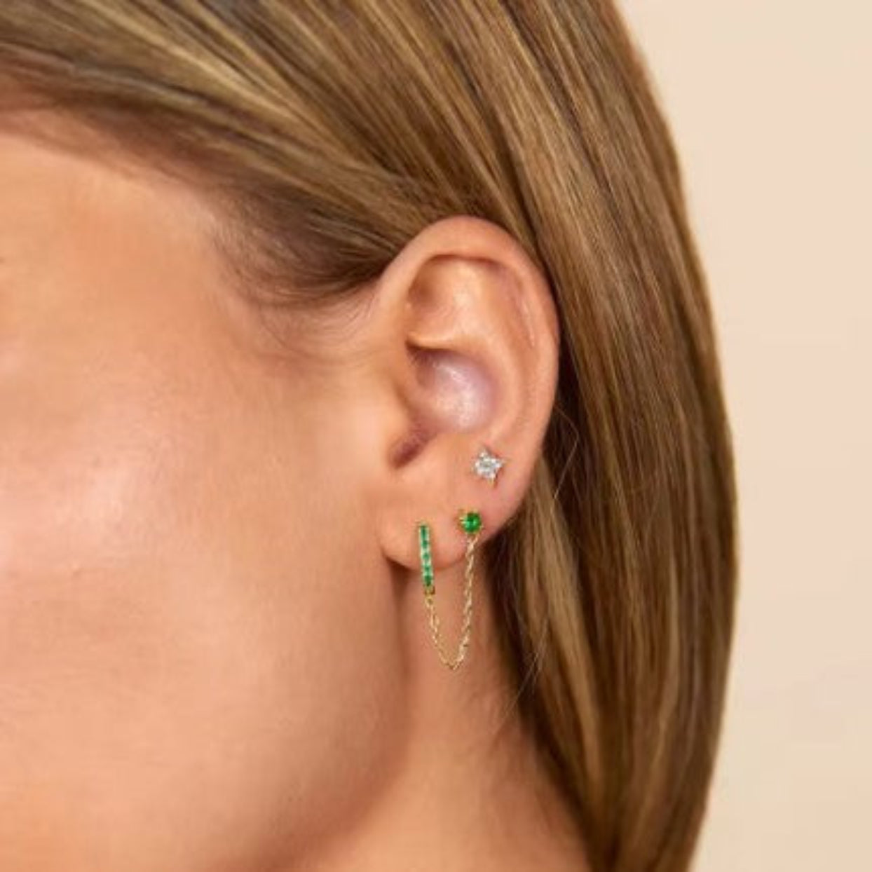 Close-up of an ear wearing gold hoop earrings with green gemstones on a beige background
