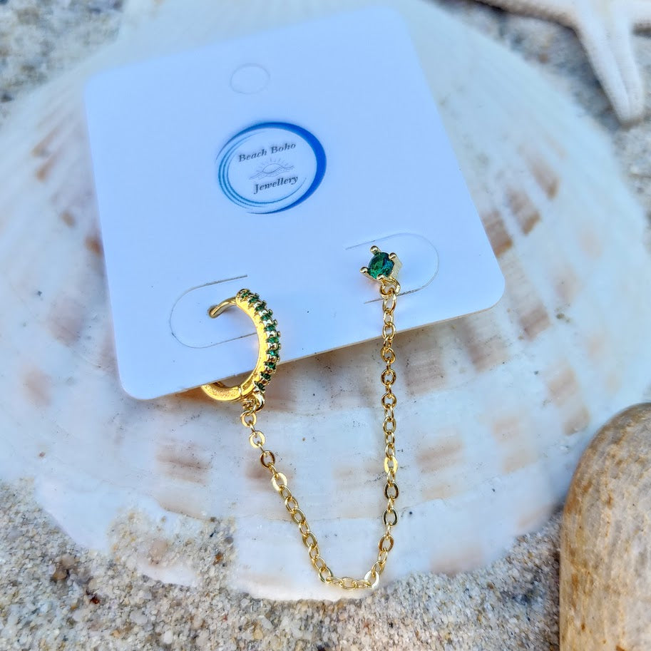 Gold earrings with green gemstones on a white card with 'Beach Babe Jewellery' branding, placed on a shell.