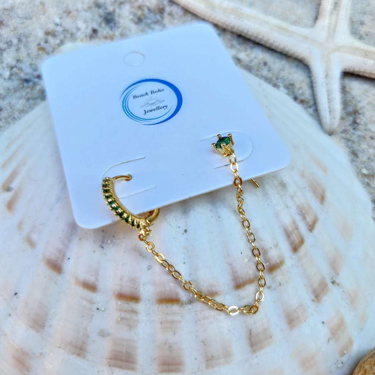 Gold chain earring with green gemstone on a branded card, placed on a shell.