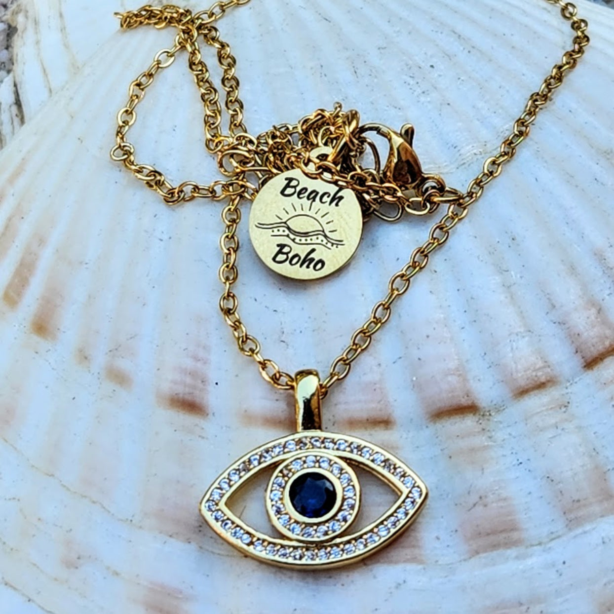 Gold necklace with eye-shaped pendant and 'Beach Boho' charm on a shell background