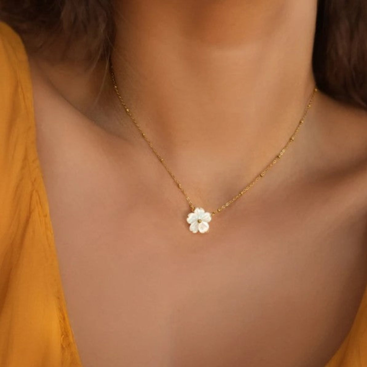 Gold necklace with a white flower pendant worn by a person in a yellow top.