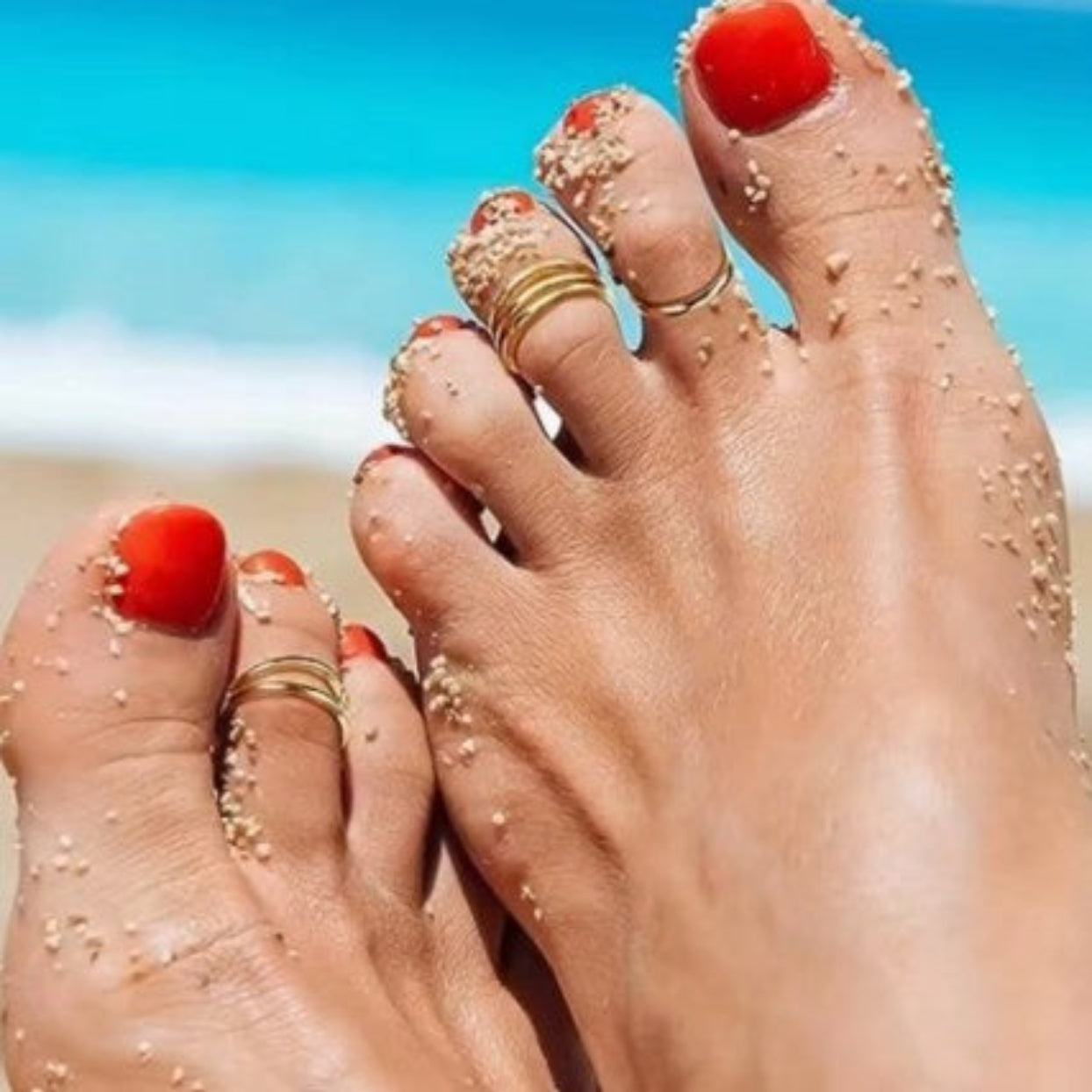 Close-up of feet with red nail polish and gold rings on a beach.