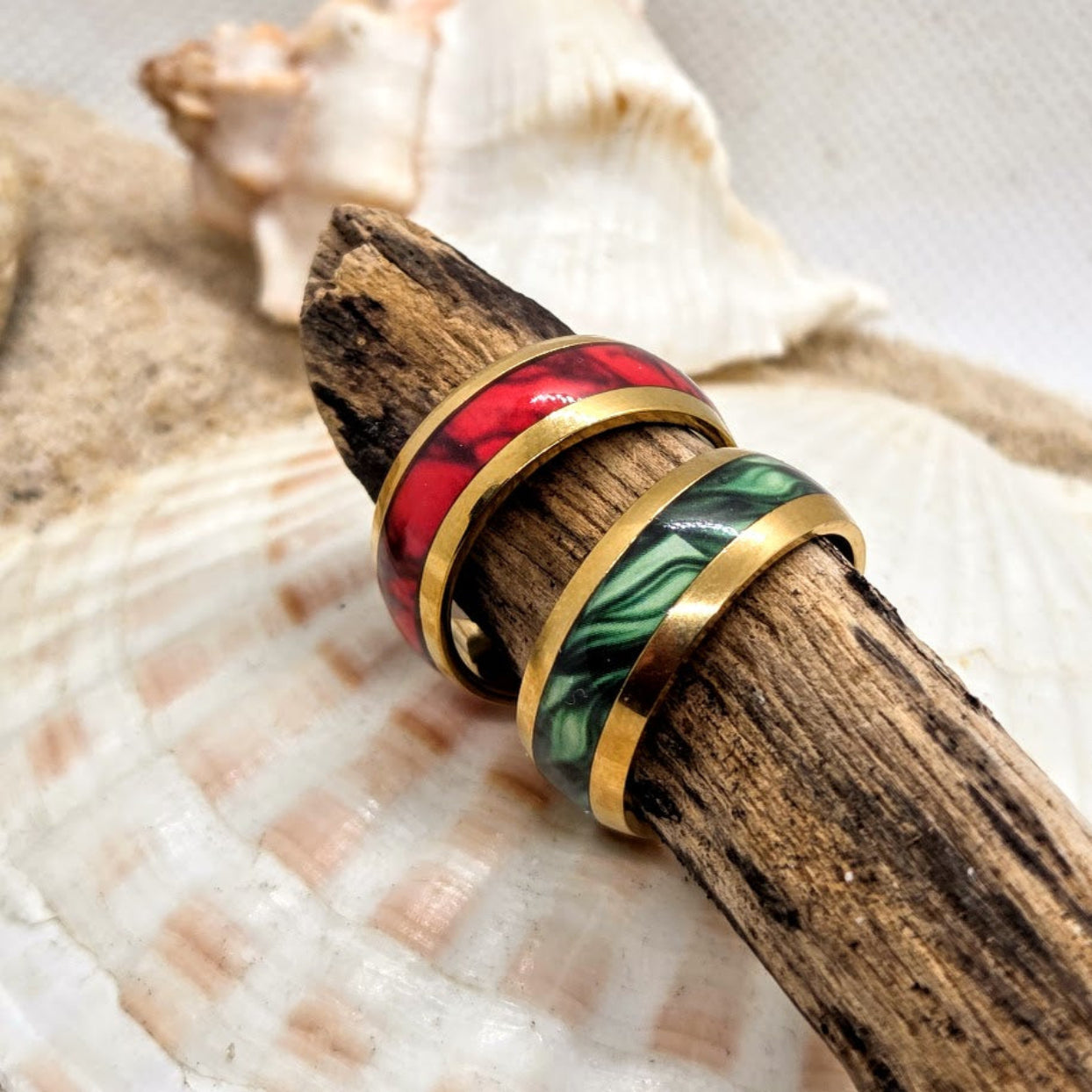 Two rings with gold bands and colorful inlays on a piece of wood with seashells in the background.