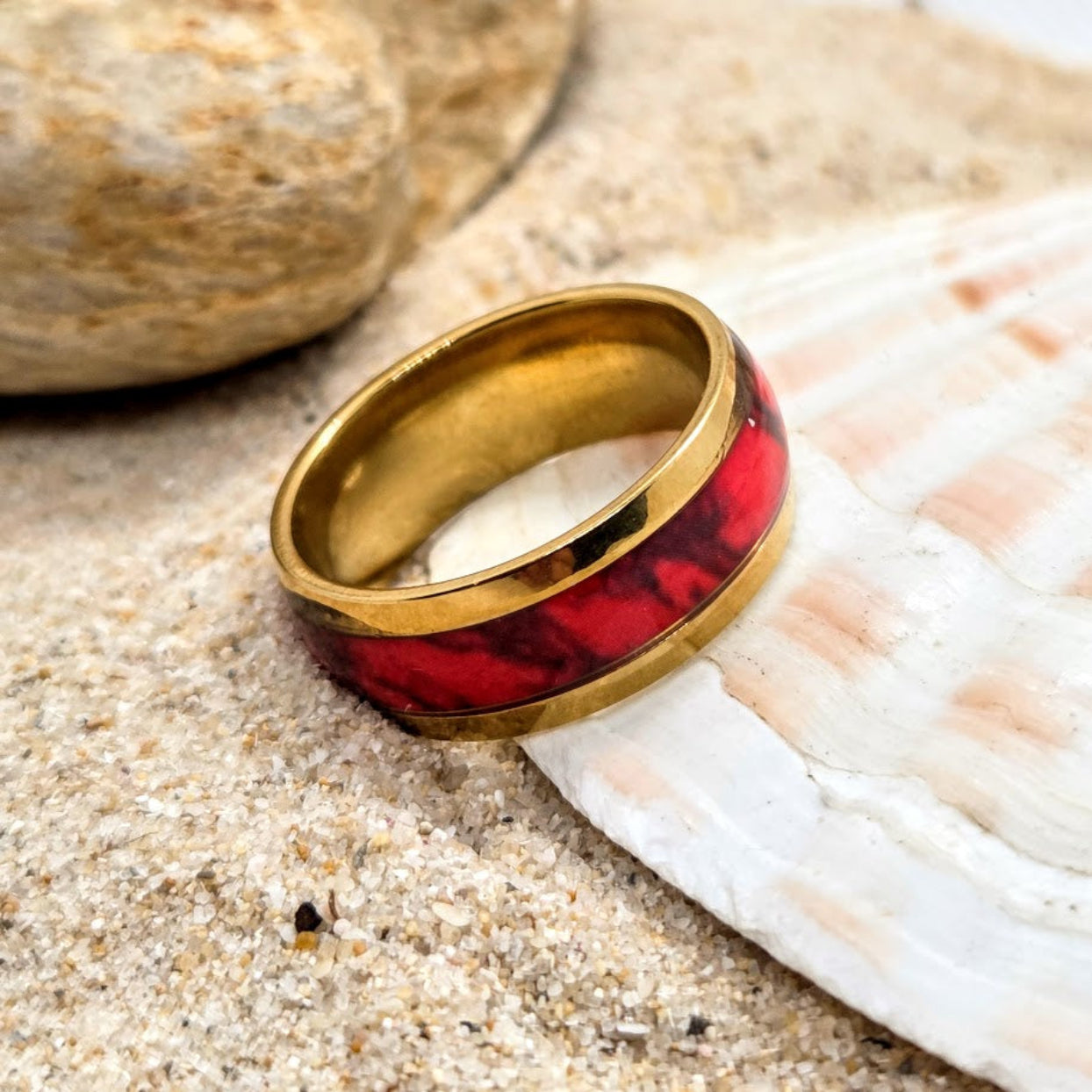 Gold and red ring on a sandy surface with a shell in the background